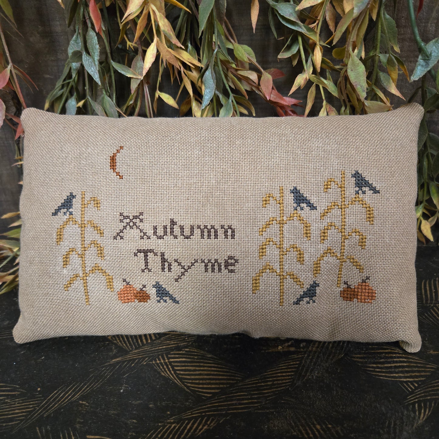 Cross-stitched pillow with 'Autumn Thyme' text and floral patterns on a textured surface.