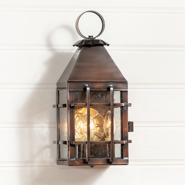 An outdoor wall light fixture with a solid antique copper finish, featuring a lantern design with visible wiring and a lit candle-like bulb within.