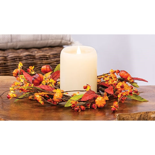 A decorative floral candle ring with a natural brown-wrapped base, adorned with colorful leaves, bittersweet branches, and orange pumpkins, surrounding a lit candle.