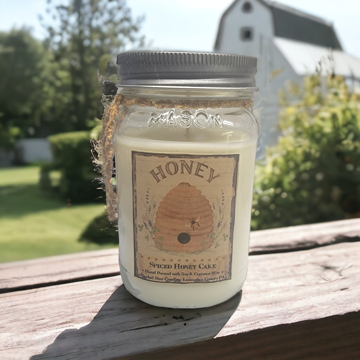 A 16 oz mason jar labeled 'HONEY' filled with a candle, set on a wooden surface with a farmhouse in the background.