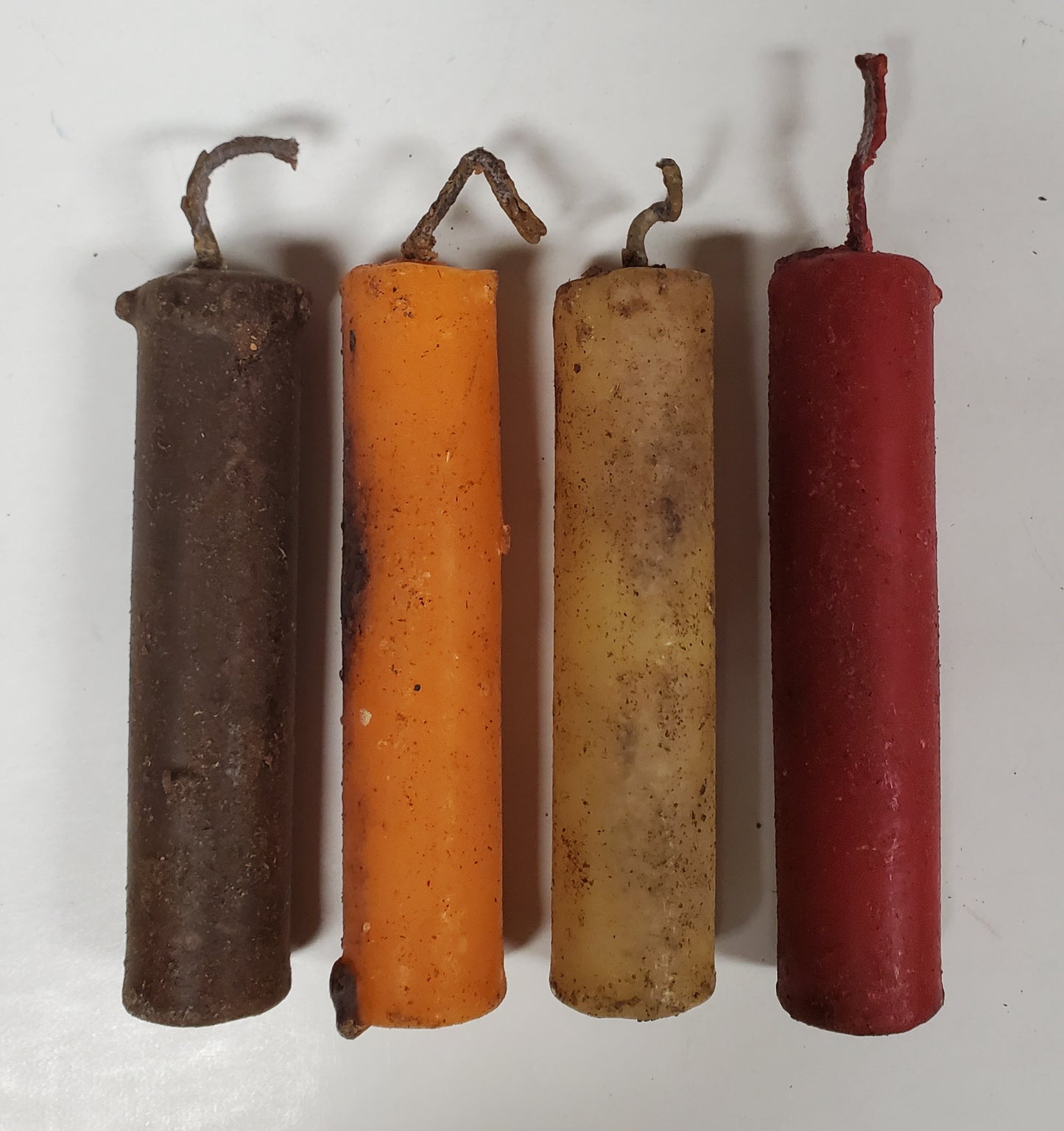 A set of four beeswax candle tapers in brown, pumpkin, ivory, and red colors, each with a primitive-looking wick and end cap.