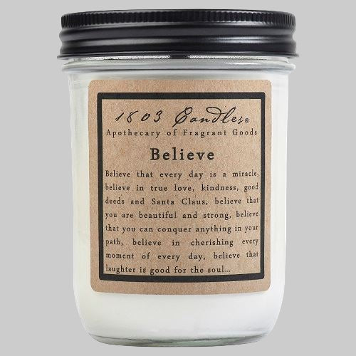 A white jar candle with a label that reads 'Believe' and has a quote underneath. The candle is placed in a clear jar with a black lid.