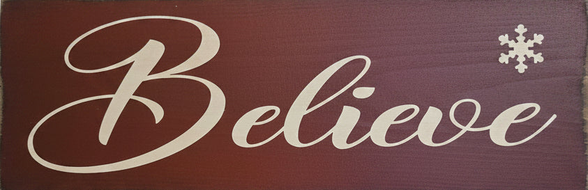 A rustic rectangular wooden sign with the word 'Believe' in large white cursive script on a dark reddish-brown background, plus a small white snowflake in the upper right corner.
