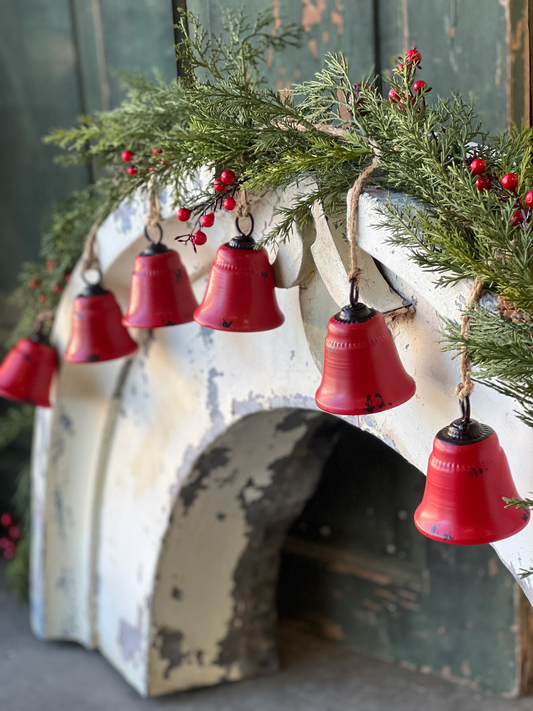 A garland of red metal bells hanging from a white structure, with greenery and red berries adding to the decor.