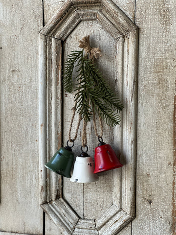 A set of three distressed metal bells in green, white, and red hanging on a white wooden wall.