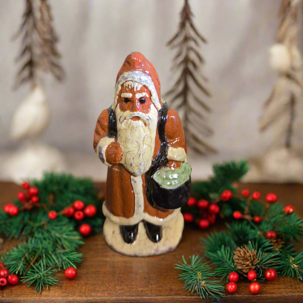 A handcrafted ceramic sculpture of Santa Claus with a red hat and beard, holding a green sack, on a wooden surface.