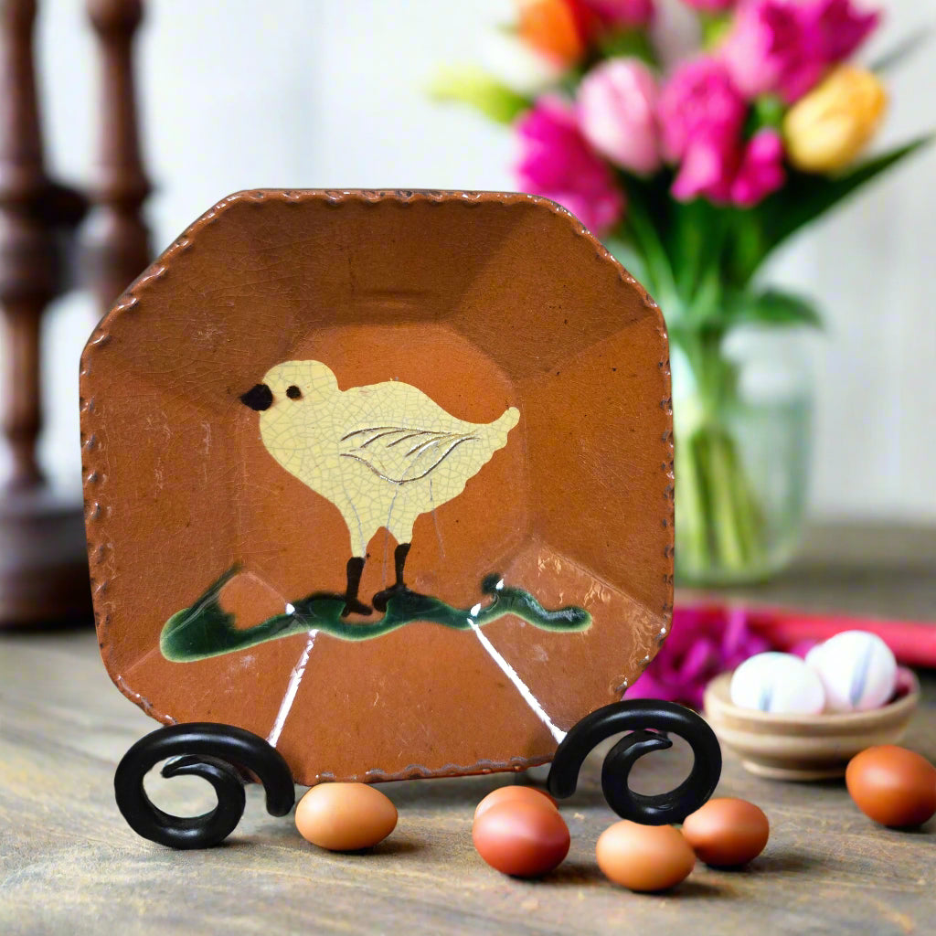 Decorative plate with a chick design on a stand, surrounded by small eggs and flowers in the background.