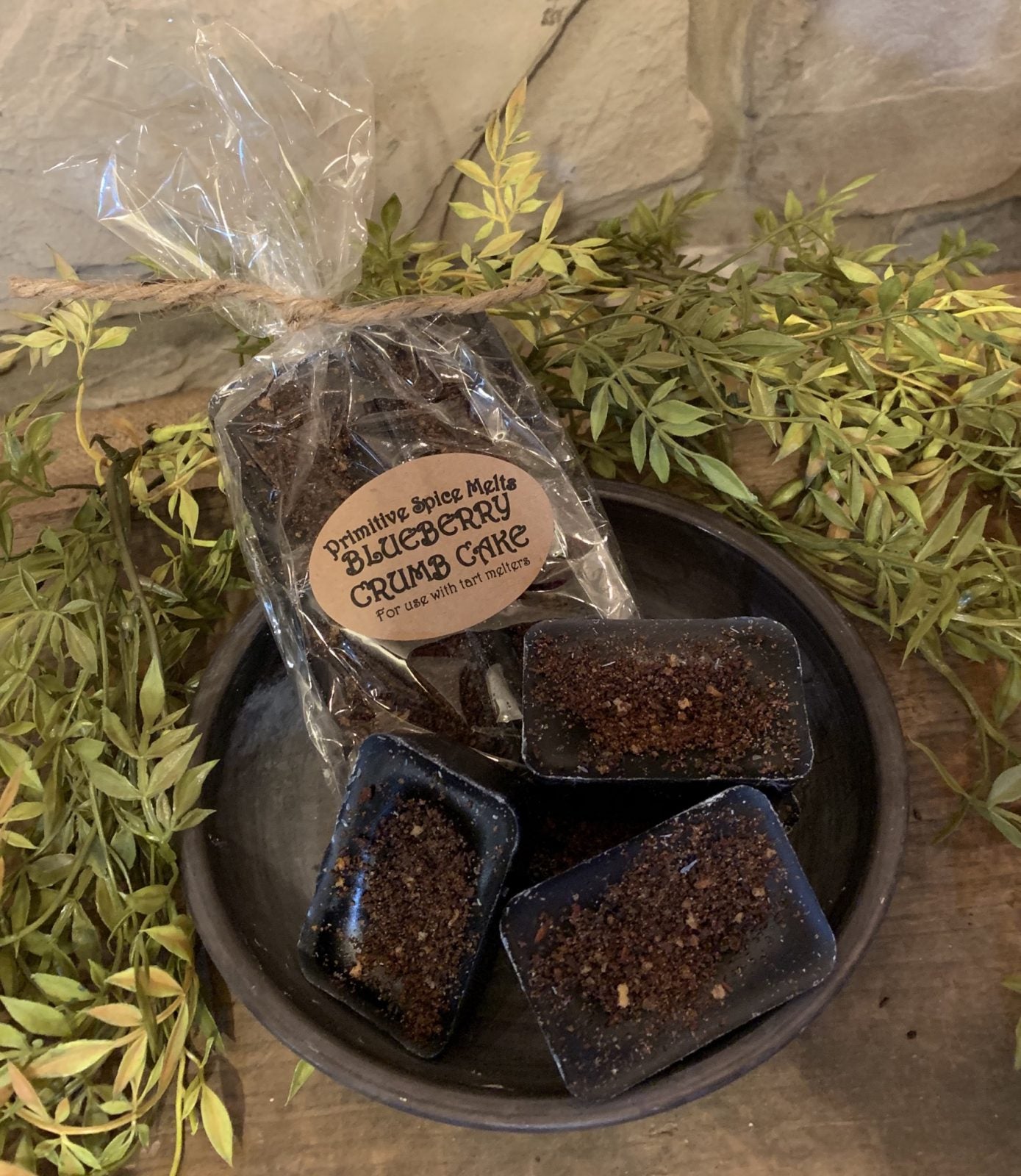 A pack of Blueberry Crumb Cake wax melts in a bowl, with a label reading 'Just Out of the Oven', surrounded by green foliage.