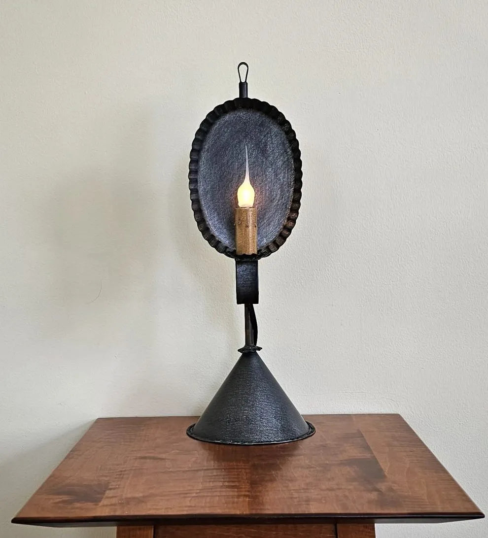 A rustic primitive style table lamp with a vintage aesthetic, featuring a conical shade on a cylindrical base, displayed against an indoor backdrop.