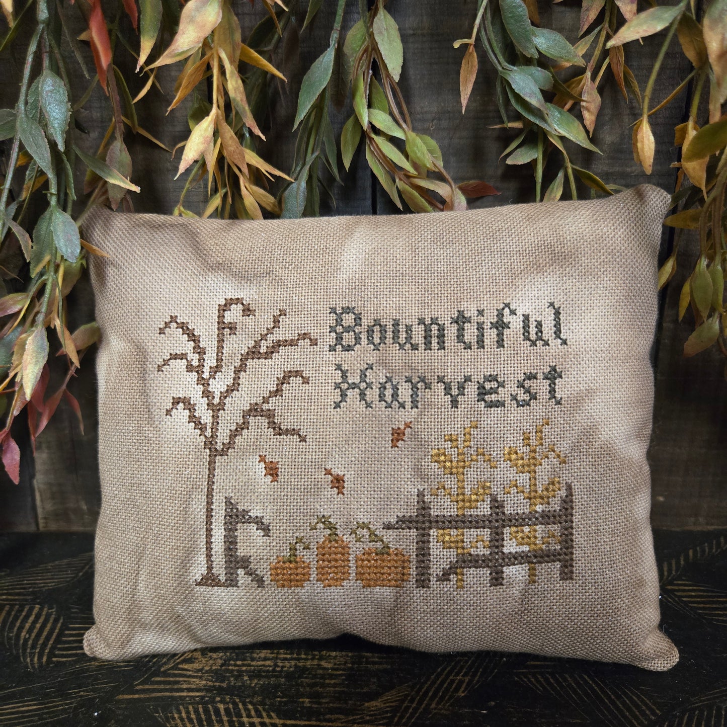 Decorative pillow with cross-stitched design and 'Bountiful Harvest' text, surrounded by greenery.