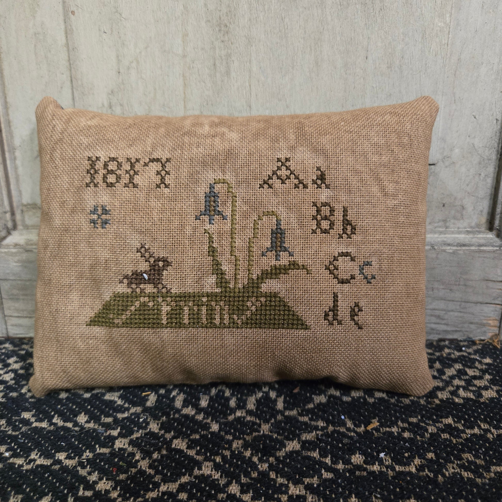 A rectangular cross stitch pillow with a bunny design and the ABC's embroidered on it, displayed on a textured surface.