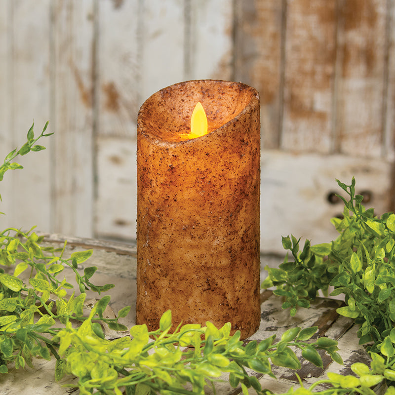 A battery operated pillar candle with a rustic wax coating in a warm burnt ivory shade, featuring a realistic flame that is lit, placed on a wooden surface with greenery in the background.