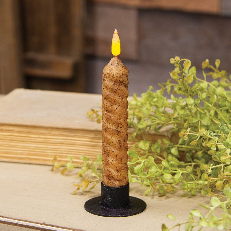 A burnt ivory vintage-style flameless candle with a twist flicker flame, placed on a black base, surrounded by greenery and a wooden object.