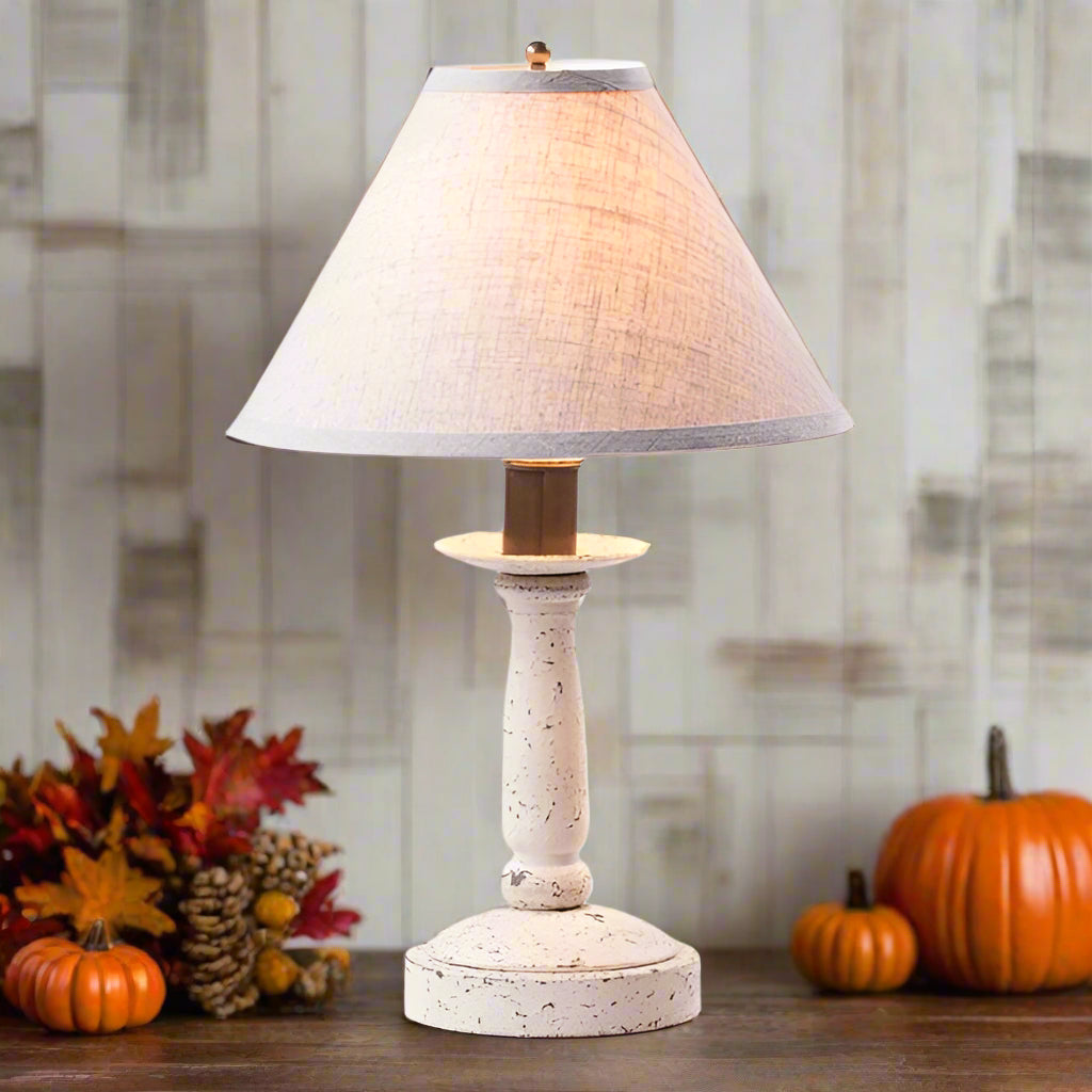 Table lamp with a white textured base and beige shade on a wooden surface with pumpkins and fall leaves.