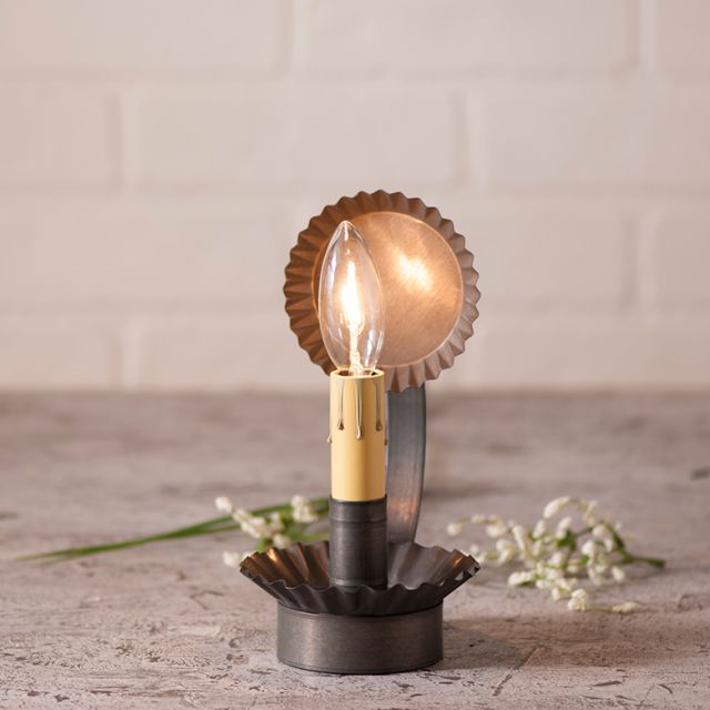 A black chamberstick lamp with a reflector, illuminated and placed on a textured surface, with a bulb gently enshrined within a circular reflector, 7 inches high.