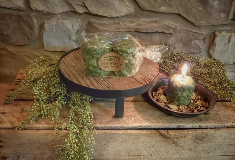 Two greenery-infused votive candles on a wooden platter with a label reading 'Cabin in the Woods', placed on a wooden surface with decorative stones and a candle lid nearby.
