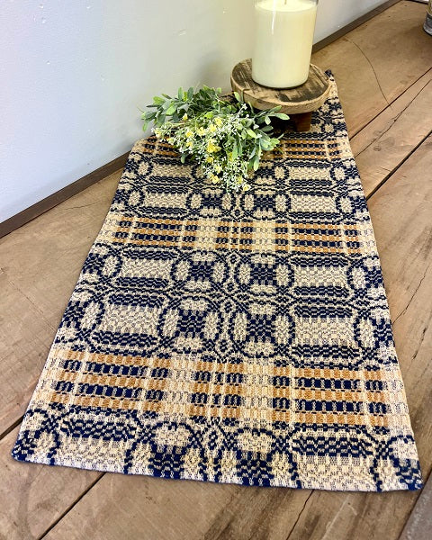 A short rectangular table runner with a geometric pattern in navy and mustard colors, displayed on a wooden surface.
