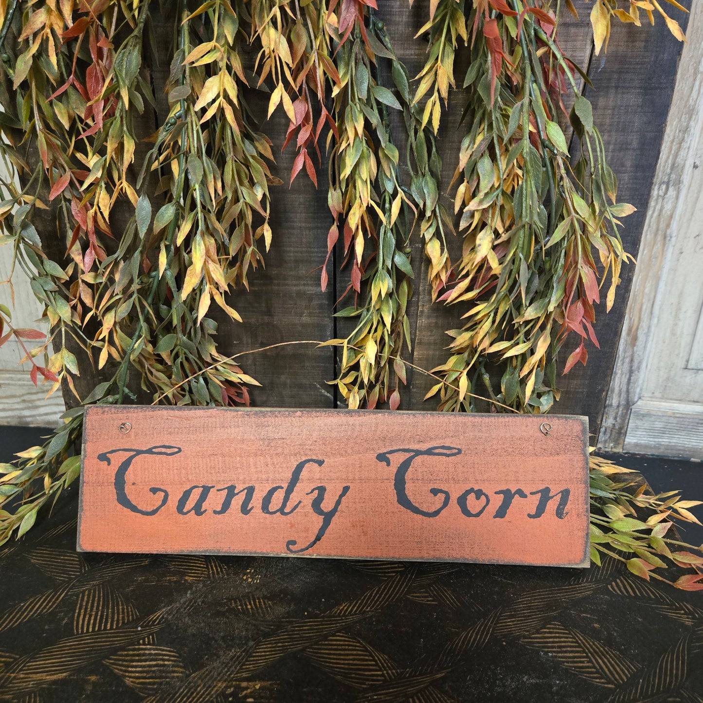 Wooden sign with 'Candy Corn' text surrounded by green foliage against a dark background