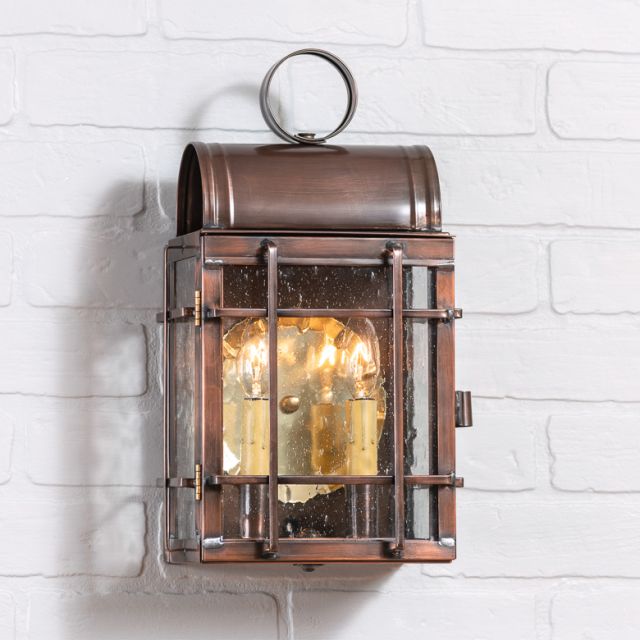 An antique copper carriage house outdoor wall light with a solid copper construction, blackened solder, and a lacquer coating, mounted on a white brick wall.