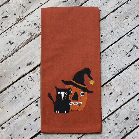 An orange hand towel with a decorative design featuring a black cat, a pumpkin, and a witch's hat on a wooden surface.