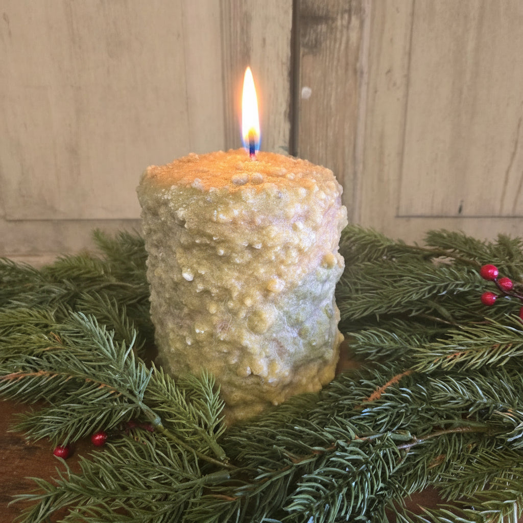 A large hearth candle with a greenish-white wax, possibly cedar and berry scented, placed on a wooden surface with green pine branches and red berries around it.