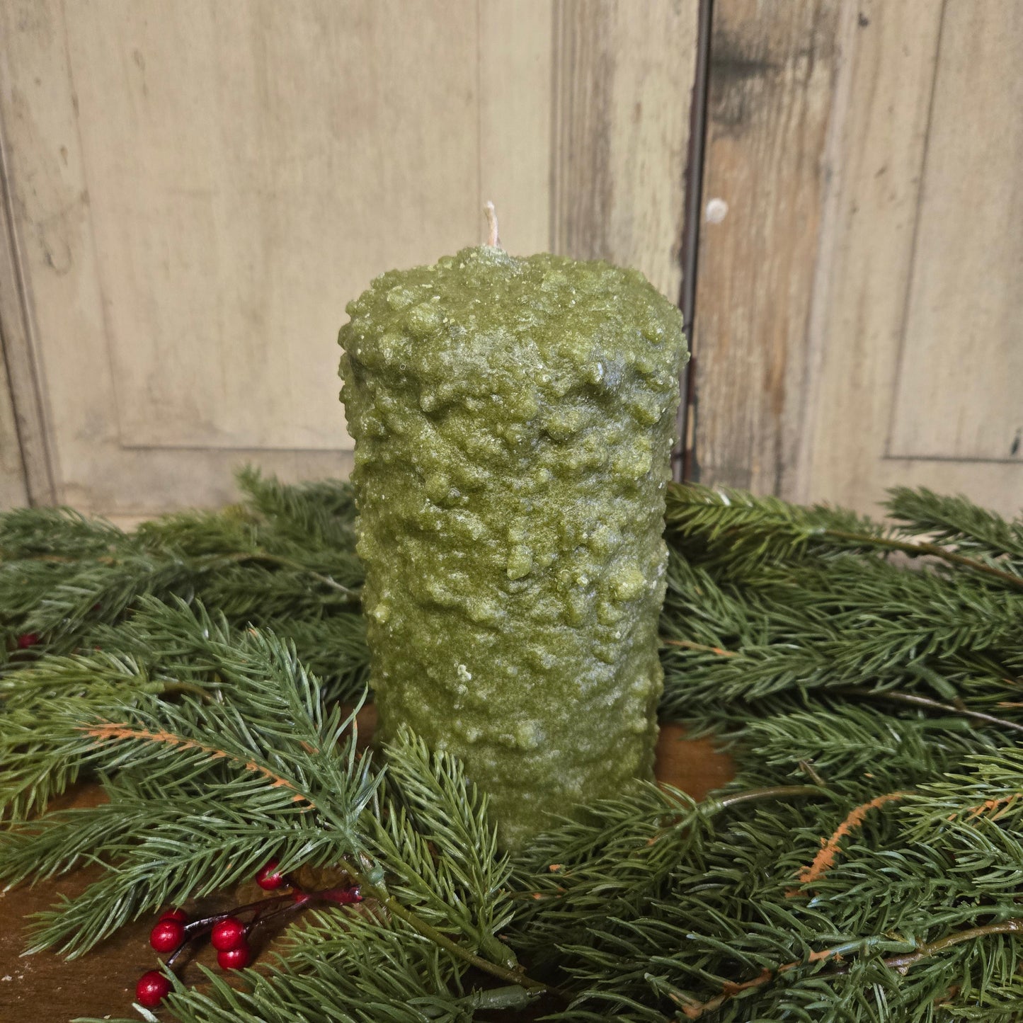 A large pillar candle with a green wax coating and a cranberry center, placed on a surface with pine branches and red berries.