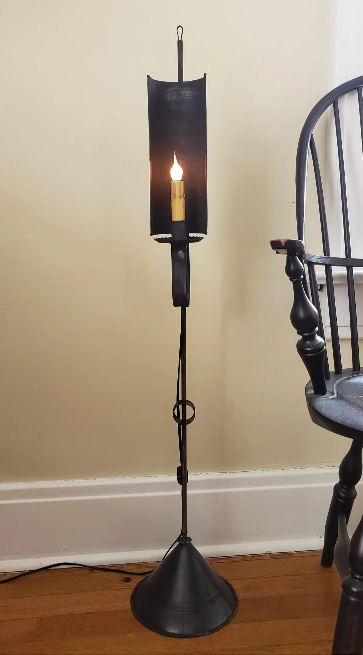 A black Charlotte Mini Floor Lamp with a primitive finish, standing in a room beside a chair with its light on, illuminating the scene.