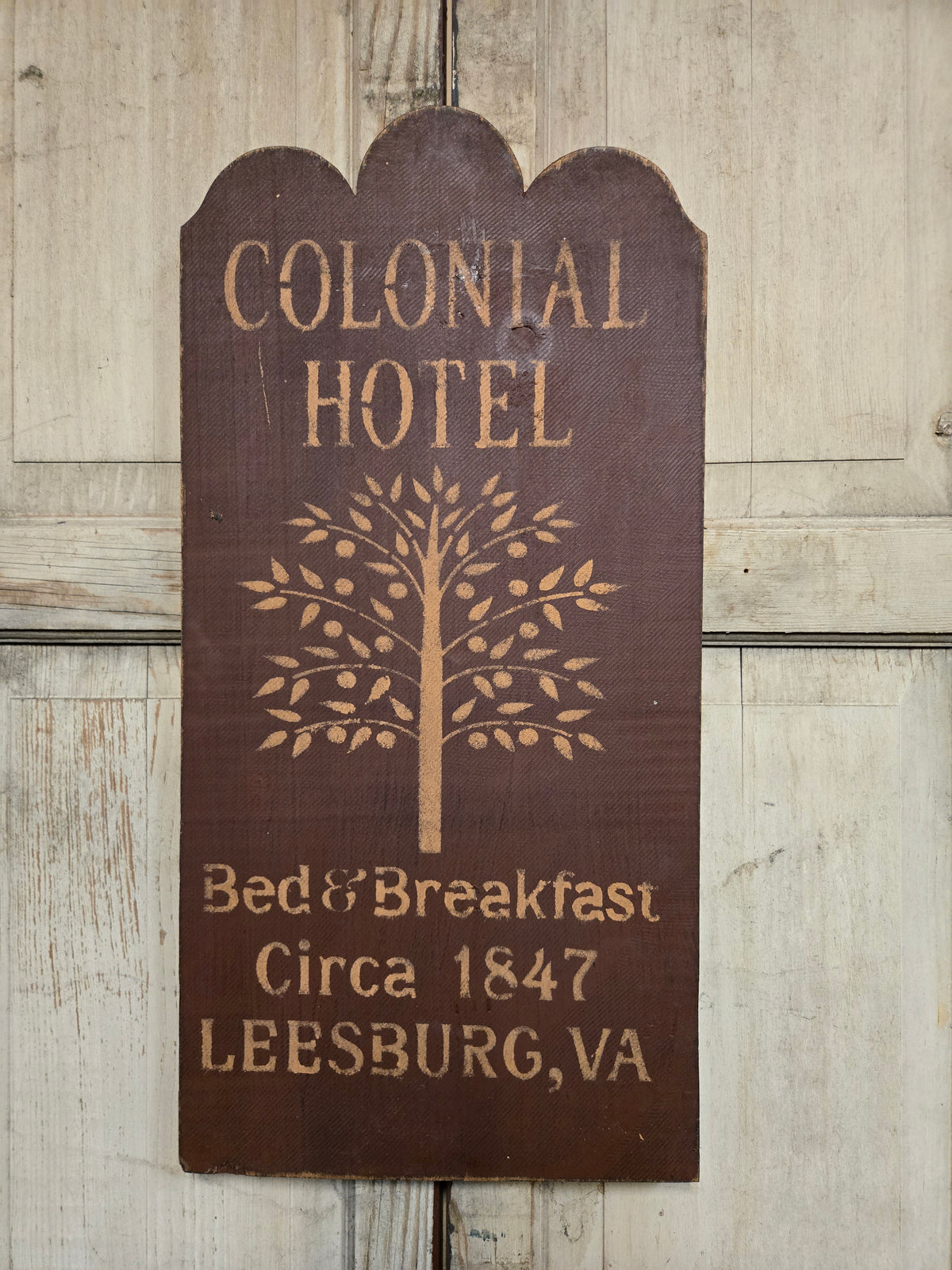A rustic dark brown wooden sign with scalloped top and engraved text reading 'COLONIAL HOTEL Bed & Breakfast Circa 1847 LEESBURG,VA' plus a tree design, shown against a light weathered wood background.