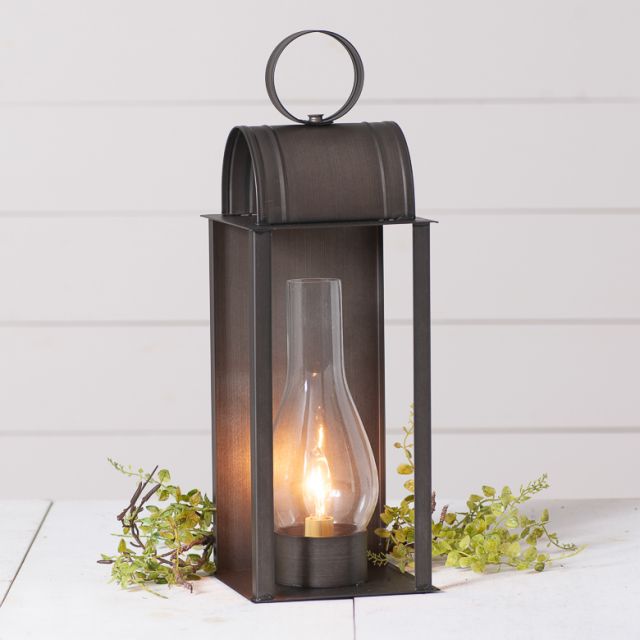 An electrified lantern light with a kettle black finish, featuring a 10-inch glass chimney, mounted on a white surface with greenery around it.