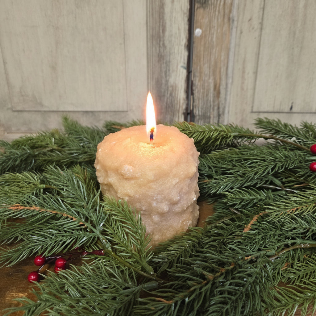 A cream colored candle with a bumpy texture surrounded by Christmas greenery