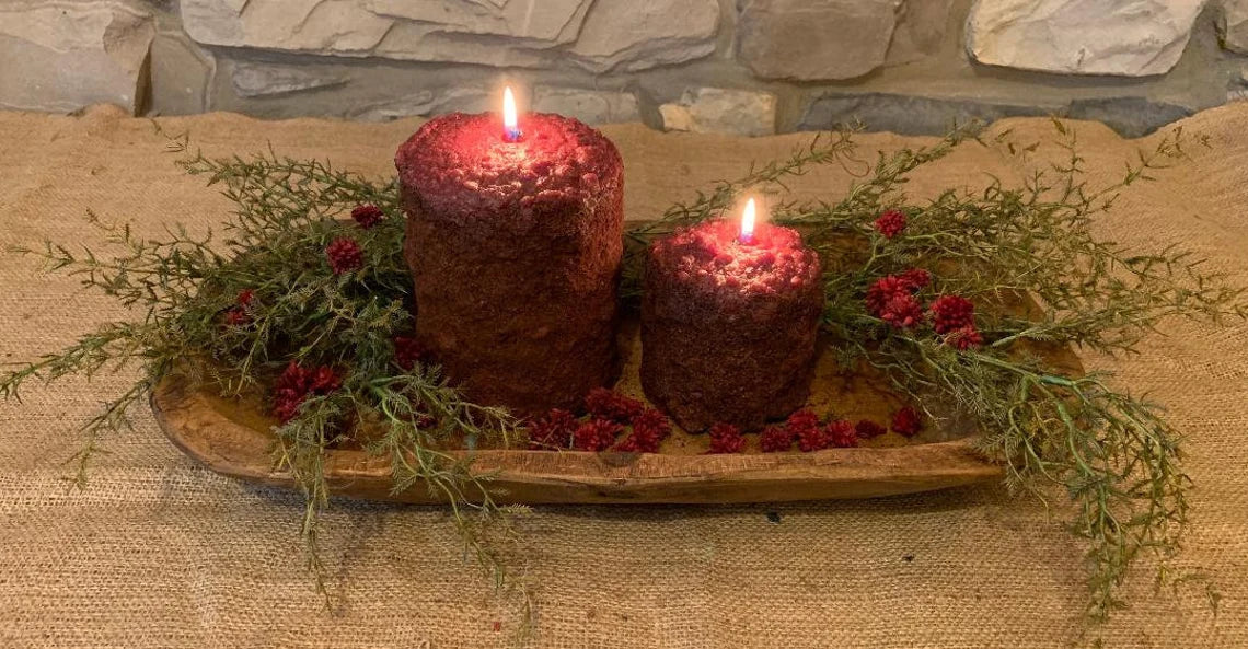 Two different size burning candles in a wood bowl with green sprigs