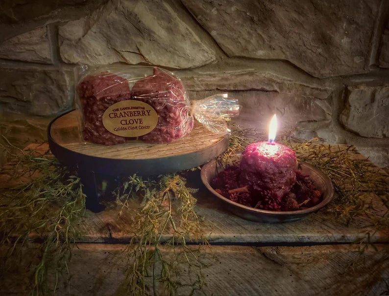 A two-pack of Cranberry Clove votive candles placed on a wooden platter, with one candle lit, surrounded by stones and a small branch on a wooden surface.