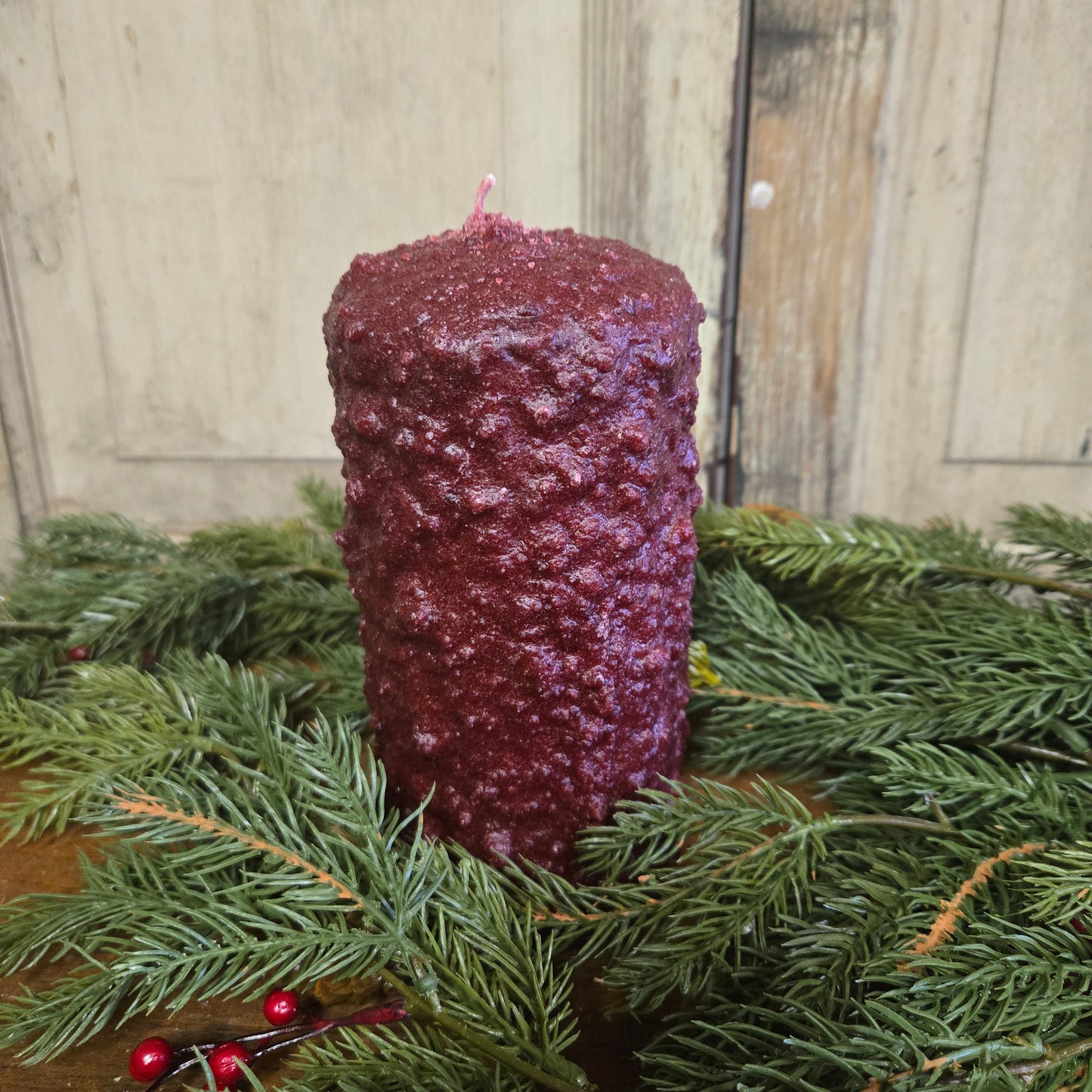A large pillar candle with a cranberry color wax, placed on a wooden surface with holly and pine branches around it.