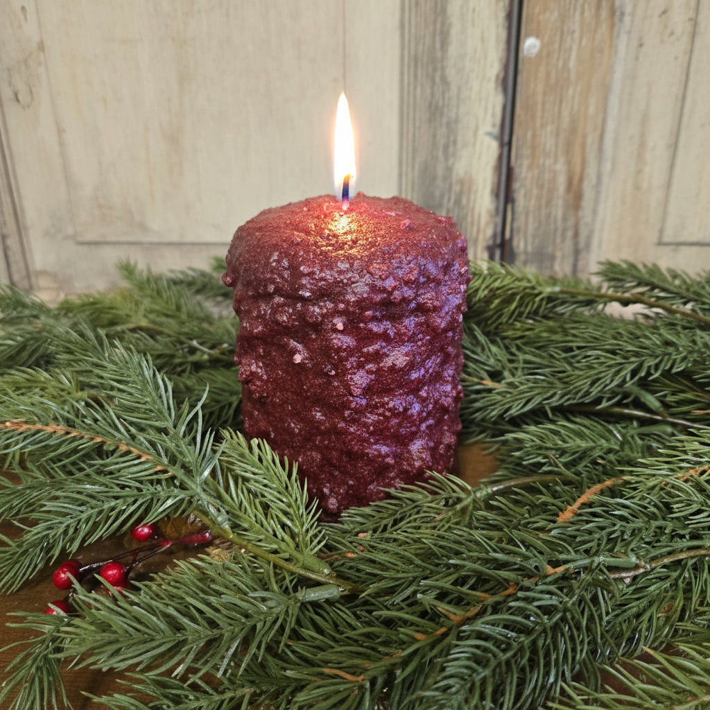 A small pillar candle with a cranberry-colored wax, placed on a pine branch, with a rustic wooden background.