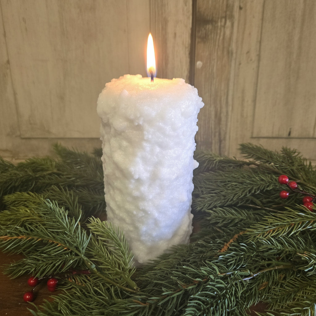 A large pillar candle with white frosting on top, surrounded by green pine branches and red berries, set against a white wooden background.
