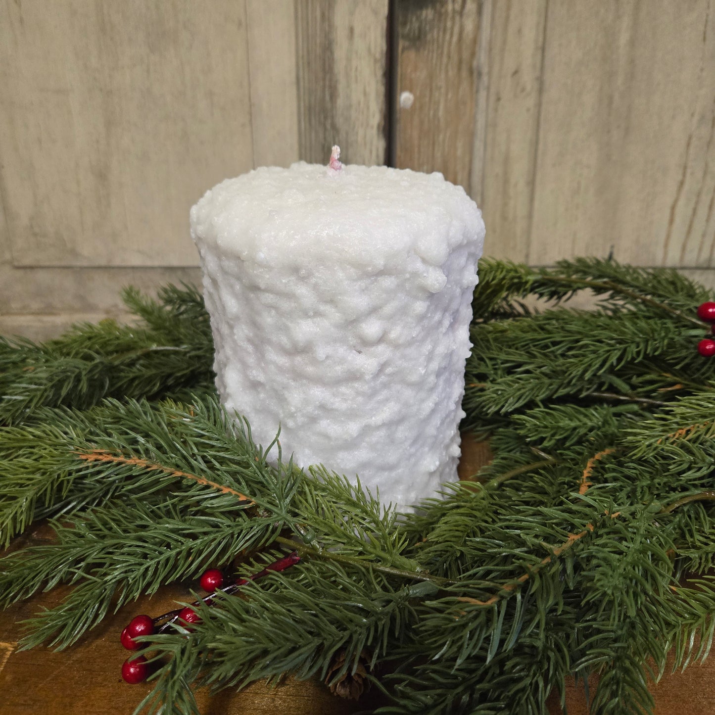A large white hearth candle with a natural wax layer, surrounded by green pine branches and red berries.