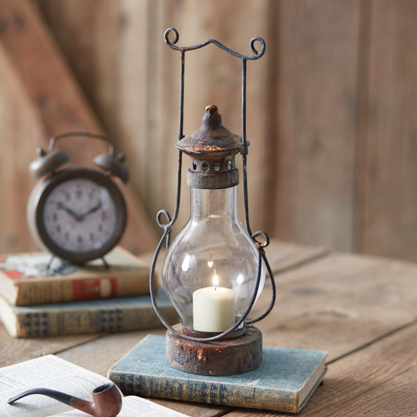 An antique-style metal and glass lantern with a lit candle inside, placed on a wooden surface next to an old clock and some books.