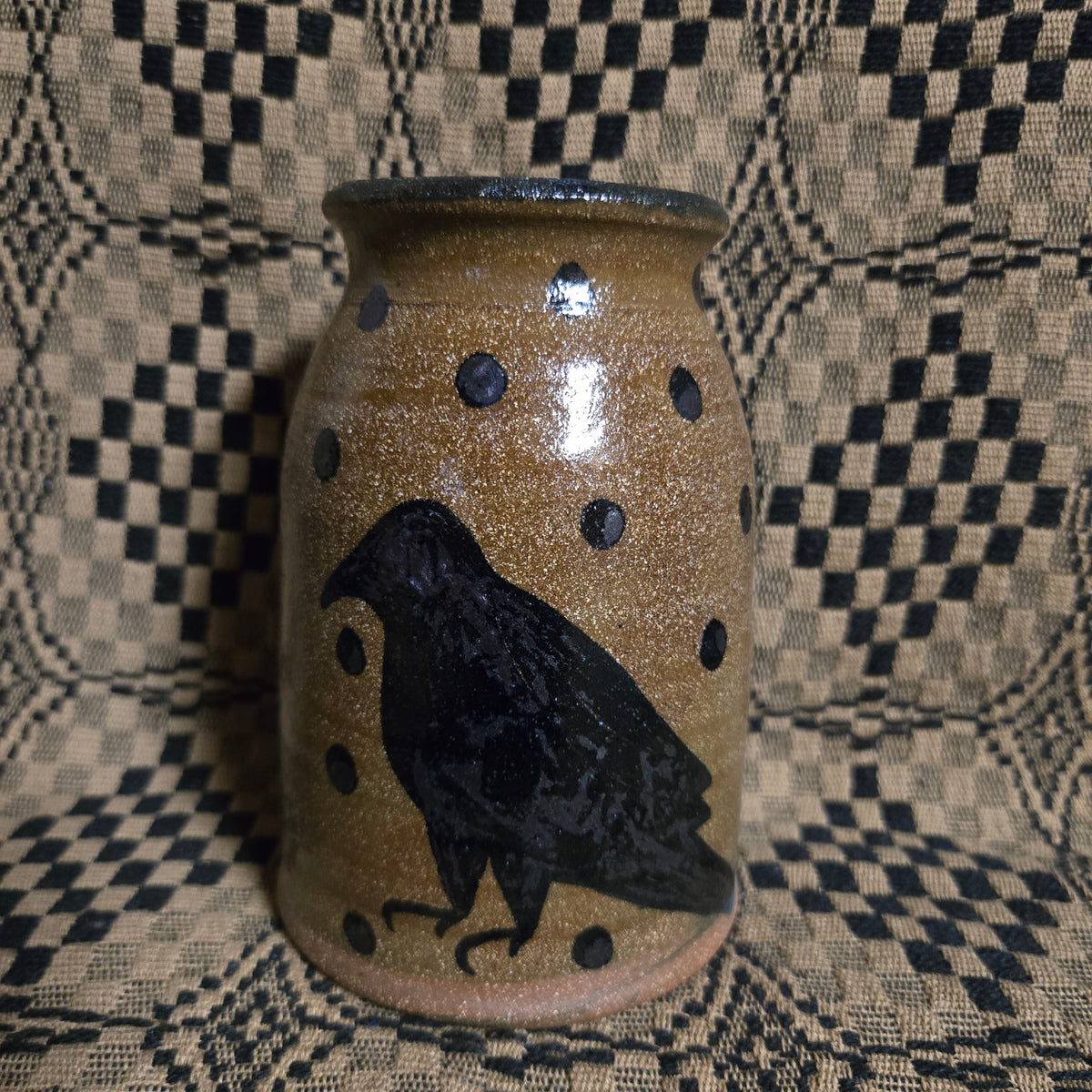 A handmade ceramic jar with a speckled brown and beige glaze, decorated with black polka dots and a black raven silhouette, set against a black and beige checkered fabric background.