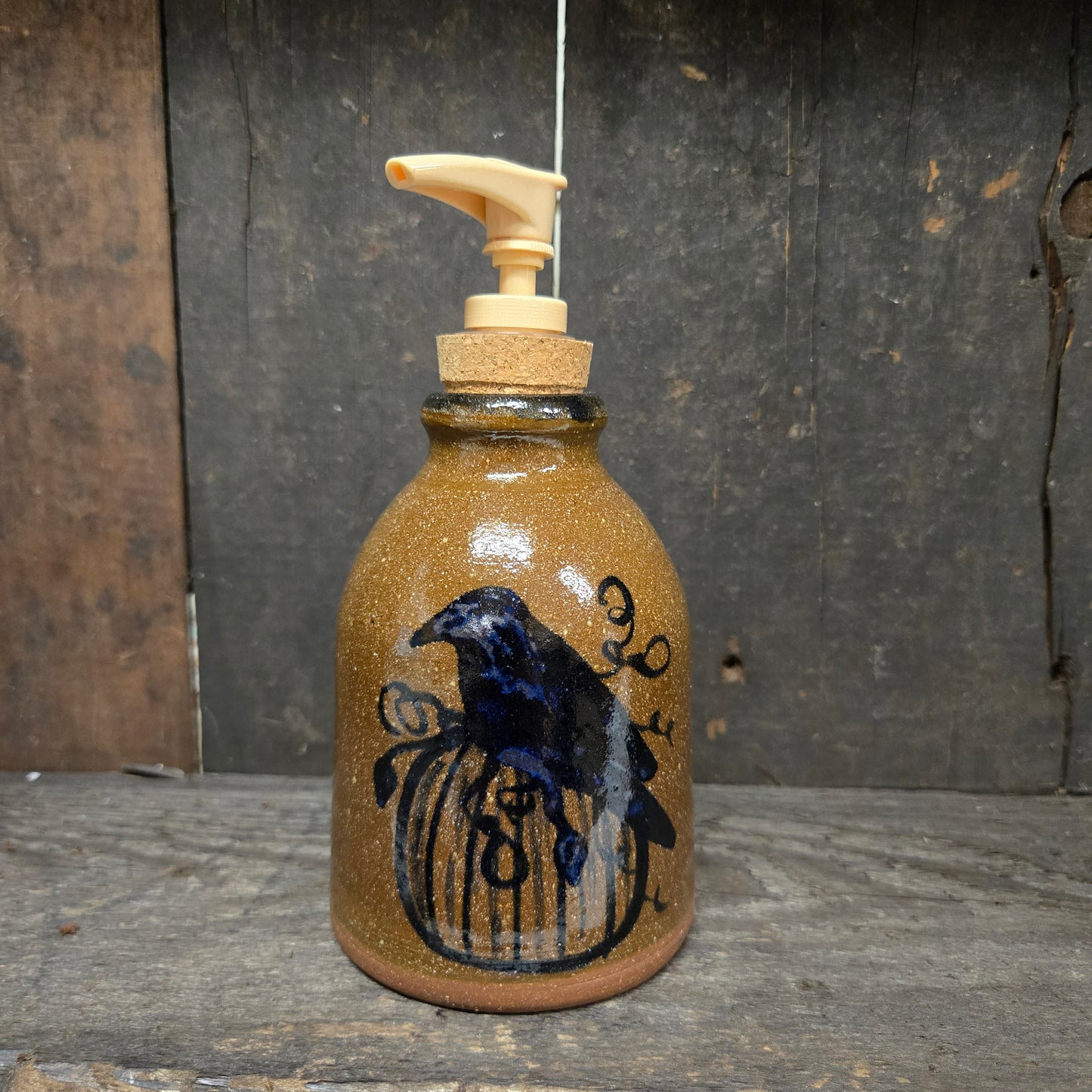 A handmade ceramic soap dispenser bottle with a brown speckled glaze, featuring a black painted raven perched on a birdcage design. The bottle has a cork stopper and a cream-colored plastic pump dispenser, set against a rustic dark wood background.