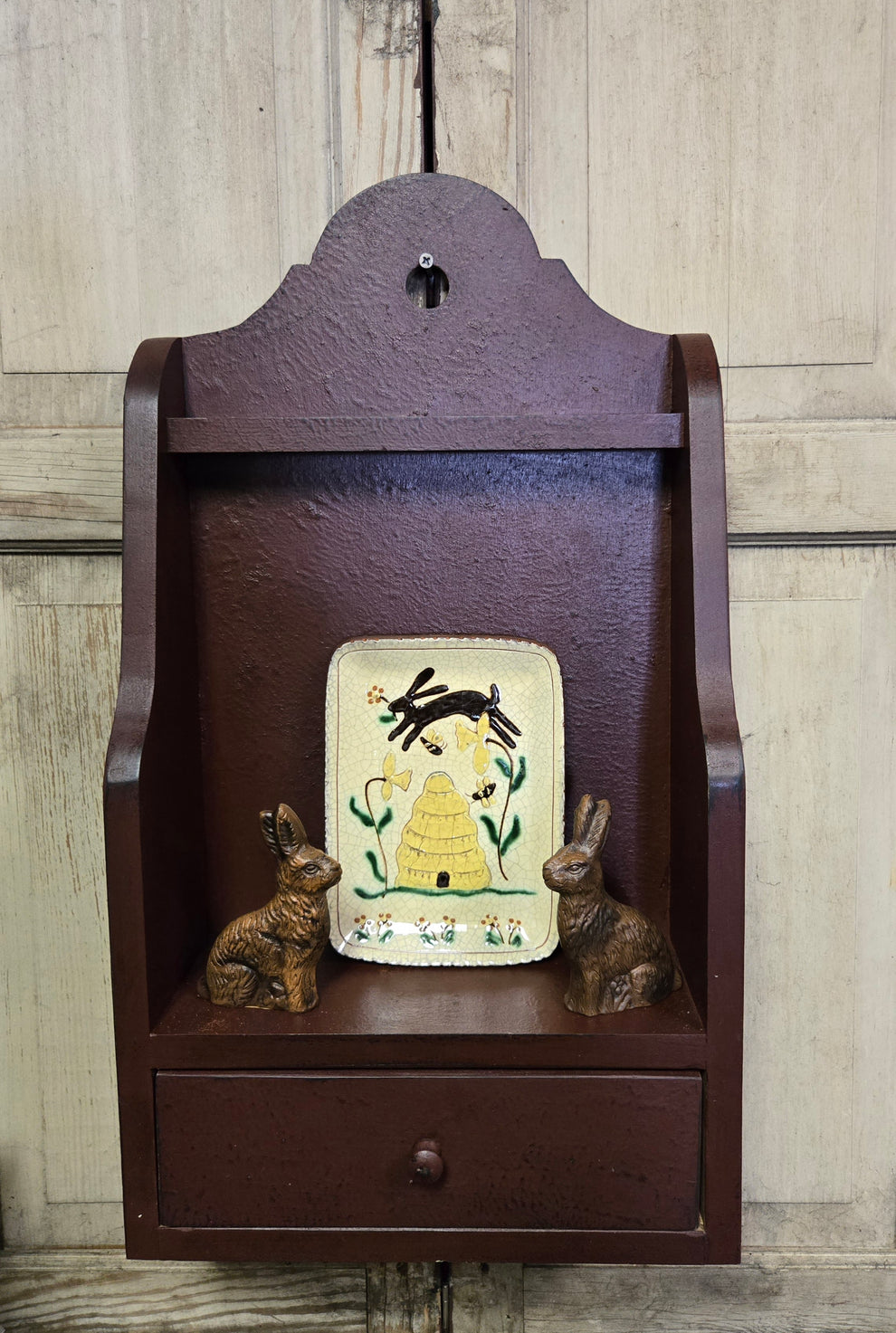 Small wooden shelf with a decorative panel and two small figurines against a wooden wall.