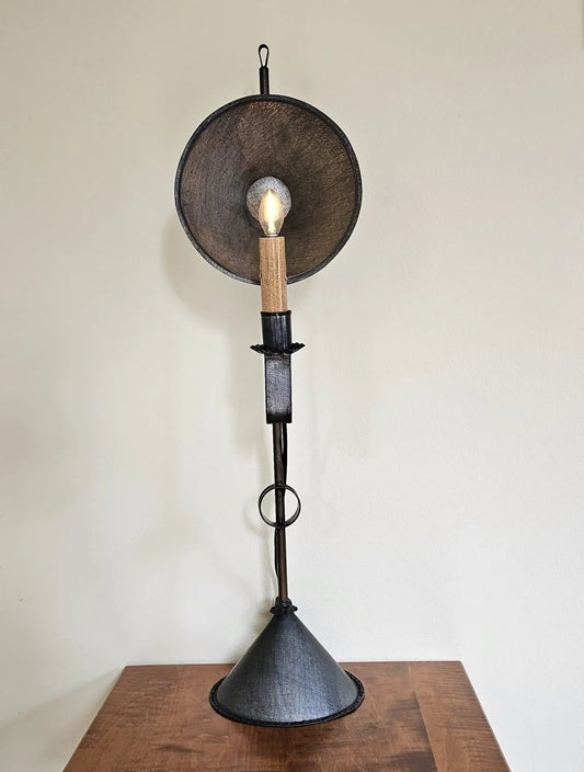 A tall table light with a primitive finish. It has a round, brown lantern-like reflector with a light bulb, mounted on a black iron base that is affixed to a wooden piece.