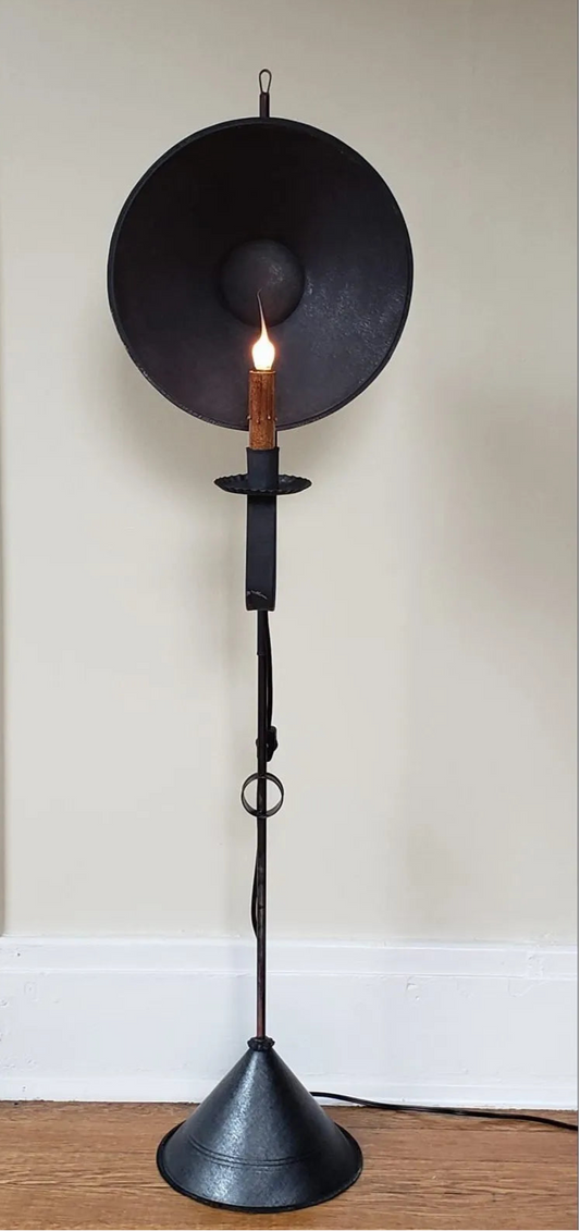 A black Cyrus Mini Tin Floor Lamp with a round tin shade and a candle-like bulb, mounted on a tripod base, placed on a wooden floor against a wall.