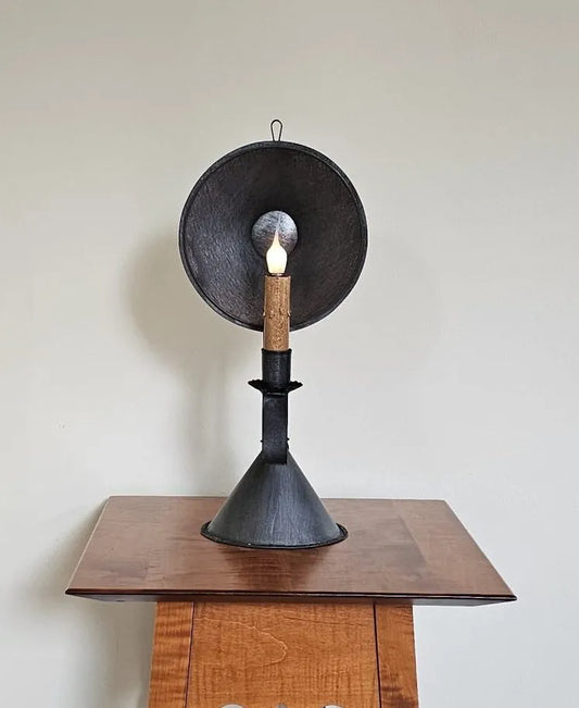 A black metal table lamp with a candle-like light source, placed on a wooden surface, against a plain wall.