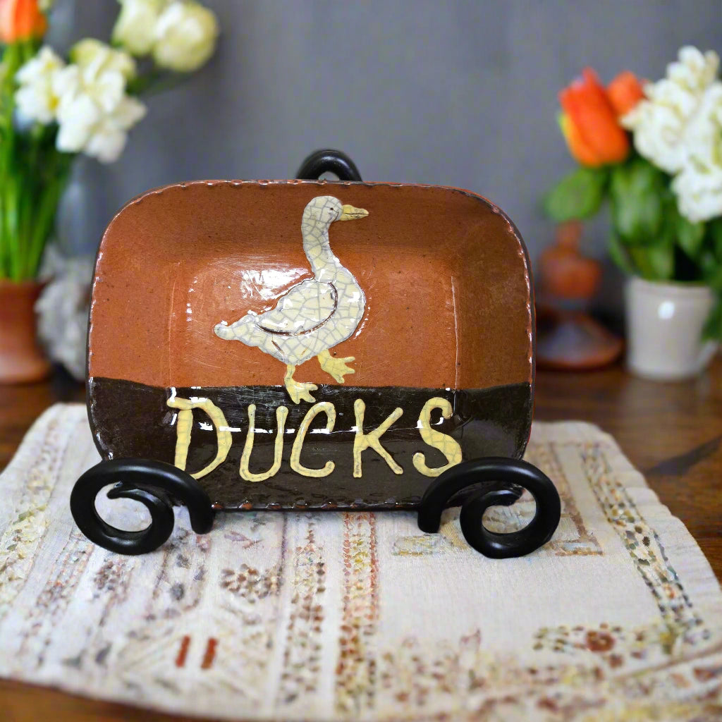 Decorative redware plate with a duck illustration and 'DUCKS' text on a table with flowers in the background.