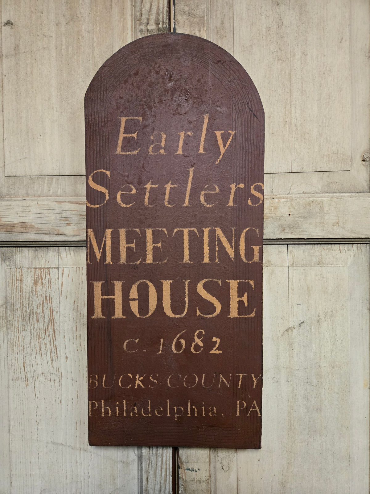 A rustic dark brown wooden sign with an arched top, featuring raised tan lettering that reads 'Early Settlers MEETING HOUSE c. 1682 BUCKS COUNTY Philadelphia, PA', displayed against a light weathered wood paneled background.
