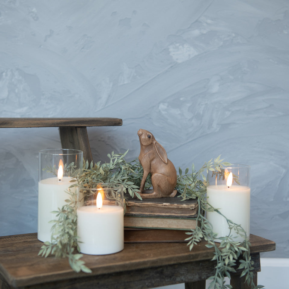 A brown resin bunny figurine placed on a wooden surface surrounded by three lit candles and decorative greenery.