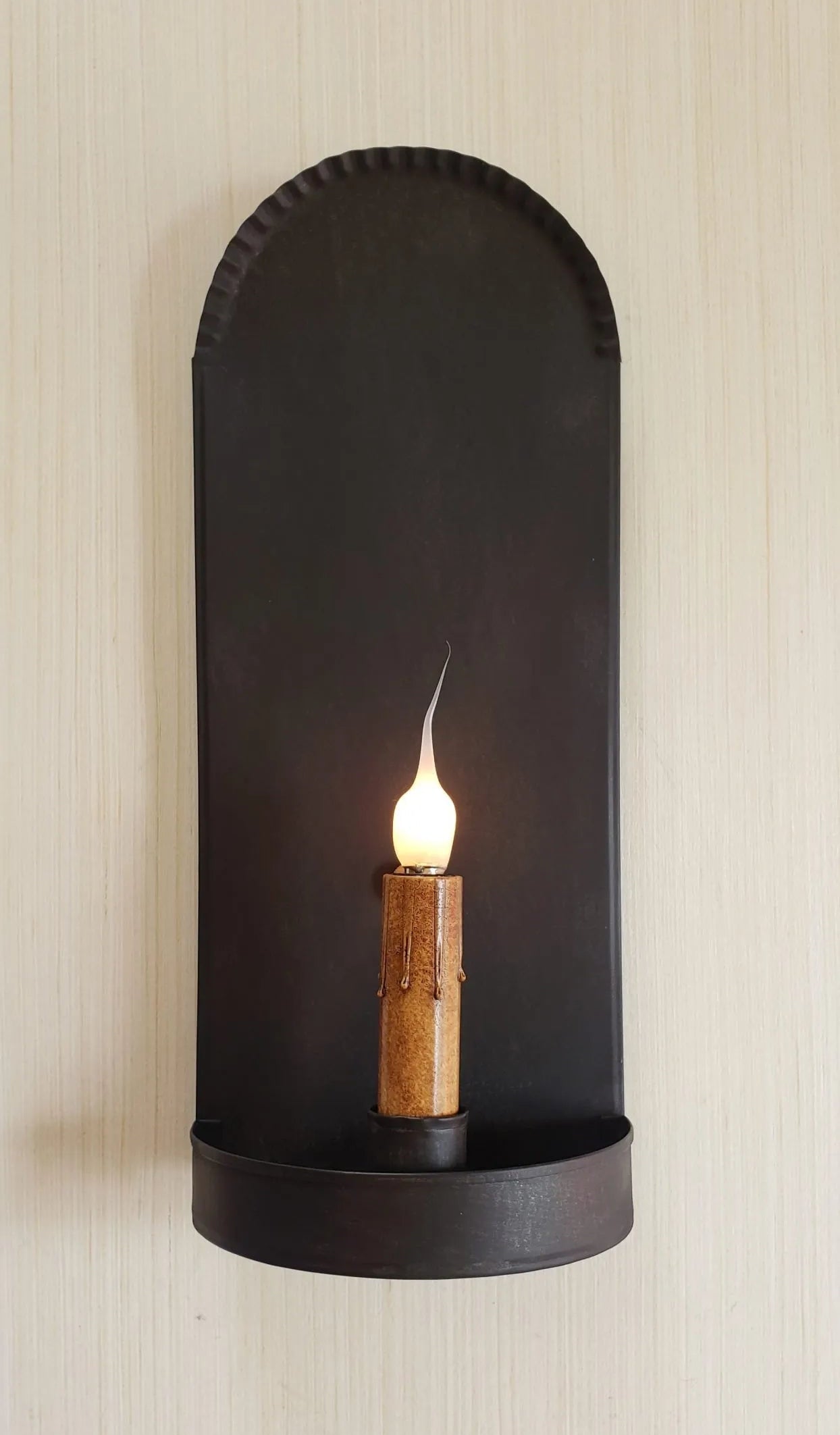 Black wall-mounted candle holder with a lit candle on a beige wall.