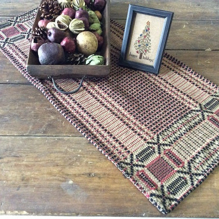 A 52 square kitchen or dining room table with a cranberry, black, and tan color scheme, featuring a geometric pattern. The table is displayed on a wooden floor next to a decorative box and a frame, with a selection of fruits and decorative items around it.