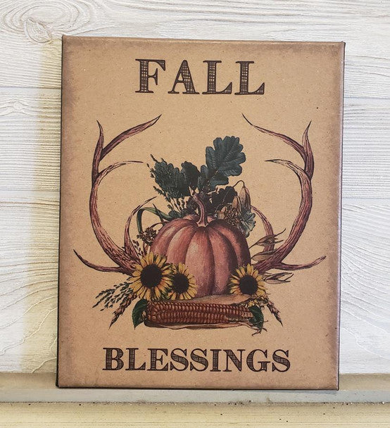 A square decorative sign with a rustic fall theme, featuring the words 'FALL BLESSINGS' and an illustration of a pumpkin, corn, sunflowers, antlers, and leaves on a tan background, displayed against a whitewashed wood wall.
