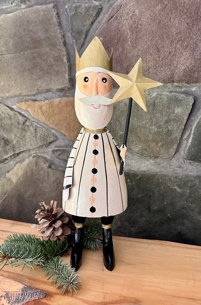 A decorative farmhouse style Santa Claus figure with a white and black striped outfit, a gold star on his head, and black boots, positioned on a wooden surface with pine cones and greenery in the background.